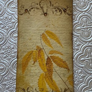 May include: A vintage-style tag with a brown ribbon, featuring a decorative floral design and a watercolour illustration of yellow leaves on a branch. The tag has a distressed, aged look and includes handwritten text.