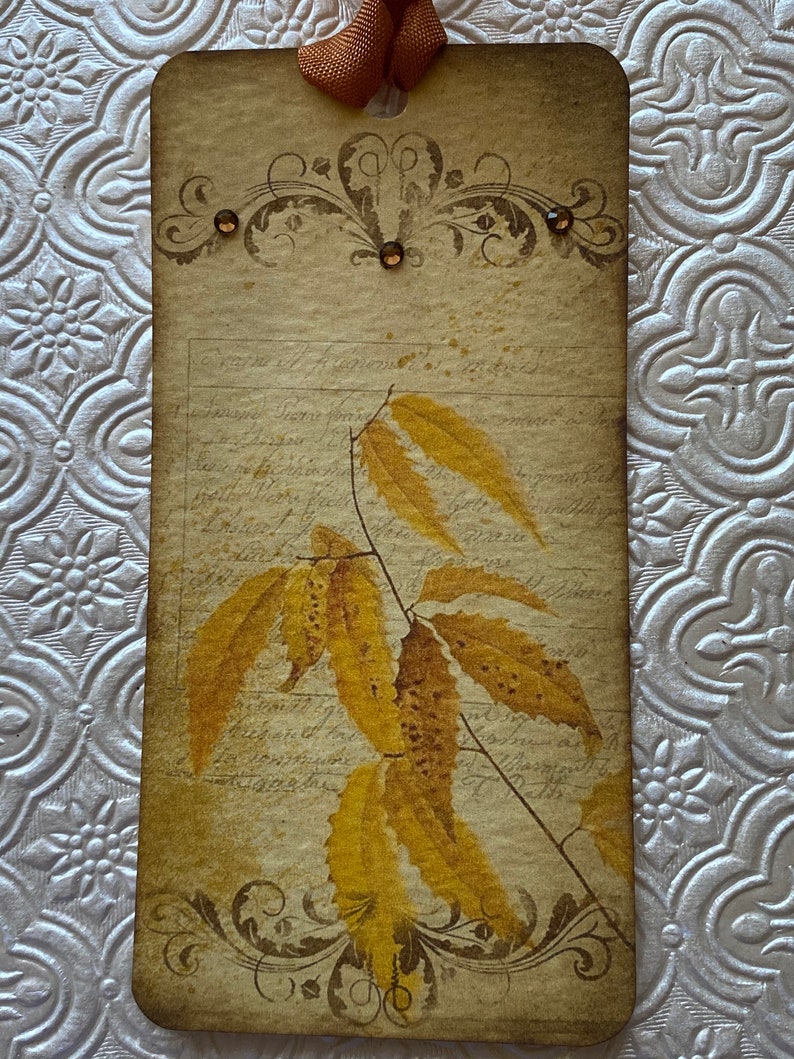May include: A vintage-style tag with a brown ribbon, featuring a decorative floral design and a watercolour illustration of yellow leaves on a branch. The tag has a distressed, aged look and includes handwritten text.