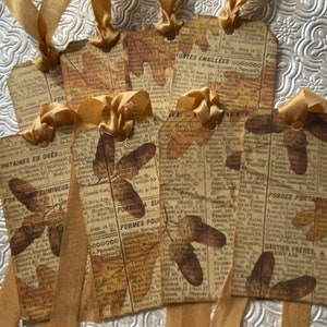 May include: Six brown paper gift tags with gold ribbon. Each tag features a vintage newspaper clipping with text and a printed image of autumn leaves. The text on the tags includes "FONTES EMAILLEES 0000000", "FORGES PORTATIVES", and "GAUTIER FRÈRES, SERVAIS."