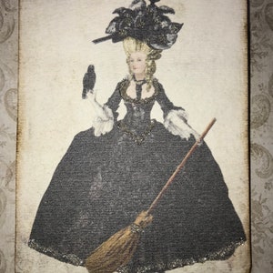 May include: Wooden tag featuring a vintage illustration of a woman in a black gown and elaborate hat, holding a broom and a raven. The tag has a black ribbon for hanging and a distressed, aged appearance.