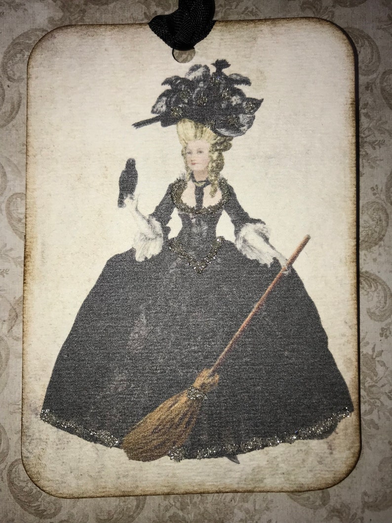 May include: Wooden tag featuring a vintage illustration of a woman in a black gown and elaborate hat, holding a broom and a raven. The tag has a black ribbon for hanging and a distressed, aged appearance.