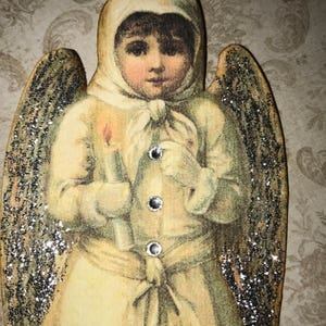 May include: A vintage-style angel ornament with silver glitter wings and a white dress. The angel is holding a candle and has a sweet expression on its face.