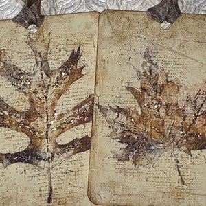 Op de afbeelding: Twee rechthoekige labels met een vintage esthetiek, met bladmotieven en glitteraccenten. De bladeren zijn in bruin- en goudtinten, tegen een achtergrond van verouderd papier met schrift. Elk label heeft een lint.