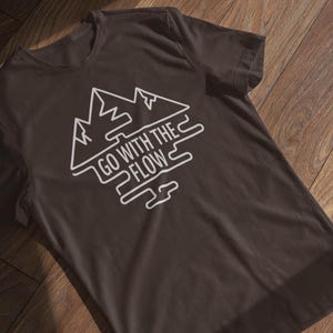 Może przedstawiać: Ciemnobrązowy t-shirt z białym nadrukiem graficznym. Projekt przedstawia szczyty gór i napis "GO WITH THE FLOW" w stylizowanej czcionce. Koszulka leży płasko na drewnianej powierzchni.