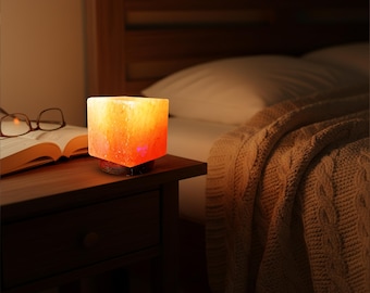 Könnte beinhalten: Eine würfelförmige Himalaya-Salzlampe leuchtet in Orange- und Gelbtönen und steht auf einem Nachttisch aus Holz. Die Lampe steht neben einem offenen Buch und einer Brille, mit einem Bett und einer Decke im Hintergrund.
