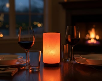 Könnte beinhalten: Eine warm orangefarbene, zylindrische Lampe steht auf einem dunklen Holztisch, flankiert von Weingläsern mit Rotwein und klaren Trinkgläsern. Im Hintergrund befinden sich ein Kamin und ein Fenster.