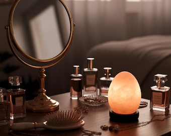 May include: A vanity table with a round, gold-framed mirror, perfume bottles, a hairbrush, and a glowing egg-shaped lamp. The lamp emits a warm, soft light, creating a cozy atmosphere. Jewelry and other accessories are also present.