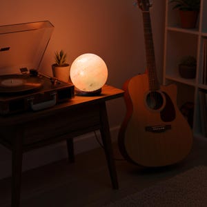 以下が含まれることがあります： 木製のサイドテーブルに、テクスチャ加工された表面を持つ光る球形のランプが置かれています。ヴィンテージのレコードプレーヤーと鉢植えの植物が近くにあります。アコースティックギターが壁に立てかけられており、温かい雰囲気を作り出しています。