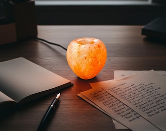 May include: A glowing orange Himalayan salt lamp shaped like a small bowl, emitting a warm light. The lamp sits on a wooden desk next to a notebook, pen, and handwritten notes. The lamp is approximately 4 inches in diameter.
