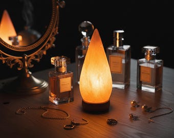 May include: A teardrop-shaped, amber-colored lamp sits on a wooden surface, surrounded by perfume bottles, jewelry, and a mirror. The lamp emits a warm glow, creating a cozy ambiance. The perfume bottles have clear glass and gold accents. Various gold necklaces, rings, and earrings are scattered around.