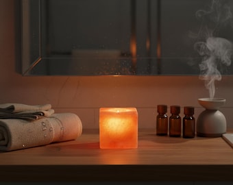 May include: A lit, square-shaped orange candle sits on a wooden surface, next to rolled towels, small brown bottles, and a white aromatherapy diffuser. Steam rises from the diffuser. A mirror is in the background.