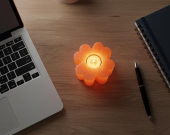 May include: A lit orange flower-shaped candle holder with a small white candle inside. The candle holder sits on a wooden desk next to a laptop, a pen, a notebook, and a small potted plant.