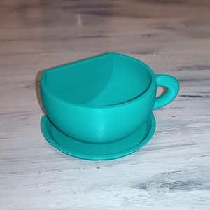 May include: A teal-colored teacup and saucer set. The teacup has a rounded shape with a handle and sits on a matching saucer. The set is made of a solid material and is suitable for decorative use.