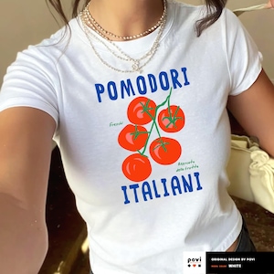May include: White t-shirt with a graphic of red tomatoes on a vine, with the words "POMODORI ITALIANI" in blue. The shirt also has the text "Freschi" and "Mercato della frutta".