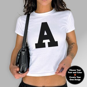 May include: A white short-sleeved t-shirt featuring a large, bold, black capital letter A printed on the center of the chest. A black leather shoulder bag hangs at the side. A circular black badge in the corner displays the text: Choose Your Font and Color, Create Your Own Design.