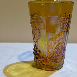 Könnte beinhalten: Ein irisierendes, bernsteinfarbenes Glas mit einem Trauben- und Blattdesign. Das Glas hat eine hohe, zylindrische Form und ist mit geprägten Trauben und Blättern verziert. Das Glas wirft einen gelben Schatten auf die weiße Oberfläche.