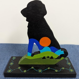 May include: A black wooden dog silhouette sculpture with a colorful landscape design. The dog is sitting on a green hill with blue and orange accents. The base is black with a teal dotted border.