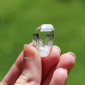 May include: A clear quartz crystal point with a natural, faceted shape. The crystal is held in a hand against a green background.
