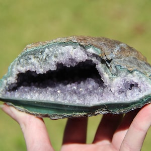 May include: A halved geode showcasing a cavity filled with sparkling, light purple amethyst crystals. The outer shell displays a mix of brown, green, and grey tones. The geode is held against a blurred green background.