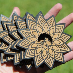 May include: A stack of six wooden coasters featuring a black and brown mandala design. The coasters are round and have a hole in the centre.