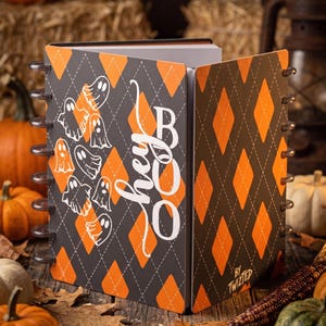 May include: A disc-bound notebook featuring a black and orange argyle pattern. The front cover displays white line-art ghosts and the text "hey BOO" in a script font. The bottom corner includes the small white text "BY TWIZTED".