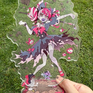 May include: A clear acrylic standee depicting an anime-style character with pink and white hair, dressed in a dark blue and black outfit with pink details. The standee has a decorative base with a pink border and a small teddy bear figure.
