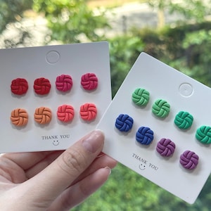 May include: A collection of colorful stud earrings in various shades of red, orange, pink, blue, green, and purple. Each earring features a knotted design. The cards read "THANK YOU" with a smiley face.