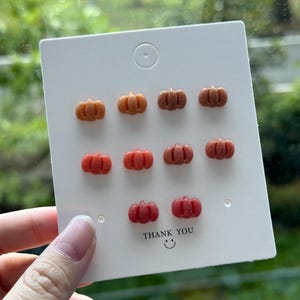 Puede incluir: Un conjunto de ocho pendientes con forma de calabaza en varios tonos de naranja y rojo. Los pendientes se muestran en una tarjeta blanca con el texto "Gracias" y una carita sonriente.