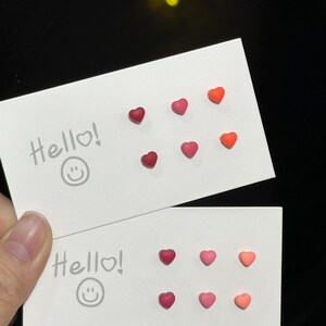 May include: Six pairs of heart-shaped earrings in various shades of pink and red. The earrings are displayed on two white cards with the word "Hello!" written on them.