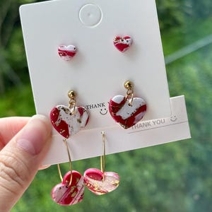 Könnte beinhalten: Ein Set aus drei herzförmigen Ohrringen mit einem rot-weiß-goldenen Marmormuster. Die Ohrringe werden auf einer weißen Karte mit dem aufgedruckten Text "Danke" präsentiert.
