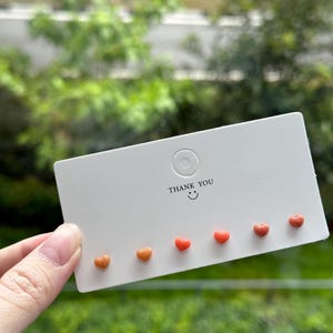 May include: A set of five heart-shaped earrings in shades of orange and red. The earrings are displayed on a white card with the text "Thank You" and a smiley face.