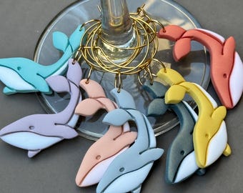 Könnte beinhalten: Eine Sammlung von bunten Wal-Wein-Anhängern. Jeder Anhänger hat eine andere Farbe, darunter Blau, Lila, Rosa, Orange, Gelb und Grau, mit weißen Akzenten. Die Anhänger sind an einem goldfarbenen Metallring befestigt.
