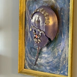 May include: A framed artwork featuring a lavender horseshoe crab adorned with sparkling embellishments. The crab is set against a textured blue and gold background, encased in a gold frame, creating a unique decorative piece.