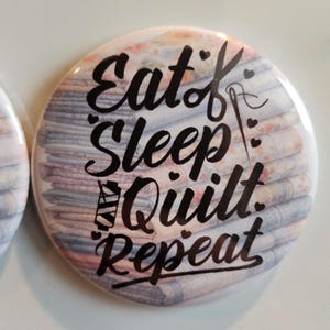 May include: A round button with a white background and black text that reads "Eat, Sleep, Quilt, Repeat". The background features a stack of colorful fabric scraps.
