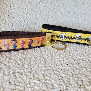 May include: Two wrist keychains featuring cartoon characters. One keychain has a brown strap with a yellow border and cartoon characters. The other keychain has a black strap with a yellow border and Snoopy.