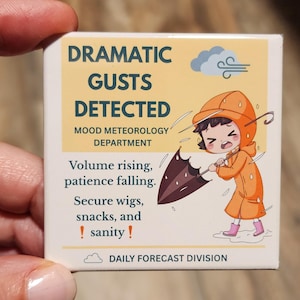 May include: A square sticker with the text "DRAMATIC GUSTS DETECTED" and a cartoon illustration of a person in an orange raincoat holding an umbrella. The sticker also includes the text "MOOD METEOROLOGY DEPARTMENT" and "DAILY FORECAST DIVISION".