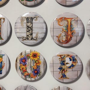 May include: A set of 10 round magnets featuring different letters of the alphabet. Each letter is surrounded by a floral design and set against a white woodgrain background.