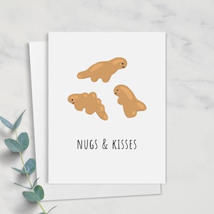 May include: A white greeting card with three cartoon dinosaur nuggets in shades of brown. The text "NUGS & KISSES" is printed in black below the nuggets. A sprig of eucalyptus is in the lower left corner.