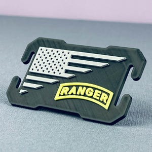 May include: A black 3D printed mask strap extender with a white and silver American flag and a yellow "RANGER" patch.