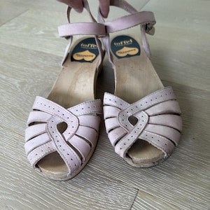 May include: A pair of light pink clogs with open-toe design. The clogs have a wooden sole and leather straps with buckle closures. The upper features a cut-out design. The brand name "toffel" is visible on the insole.