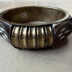 May include: A wide bangle bracelet featuring a central section of four vertical, curved brass-toned metal bars flanked by dark brown panels inlaid with small, white, oval-shaped pieces. A small white sticker inside reads MADE IN INDIA.