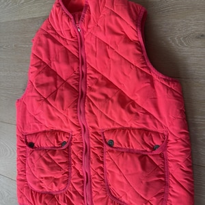 May include: A quilted, coral-colored vest with a full-length zipper and two front pockets. The vest features a diamond pattern throughout the fabric and a stand-up collar. The pockets have button closures. The vest is sleeveless.