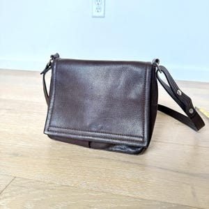 May include: A dark brown leather crossbody bag with a flap closure. The bag has a textured surface and a long, adjustable strap. The bag is approximately 10 inches wide and 8 inches tall.