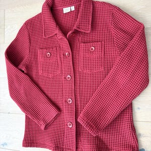 May include: A red-burgundy waffle-knit jacket with a collared neckline, two front pockets, and button closures. The jacket has long sleeves and a textured appearance. The label on the jacket is visible.