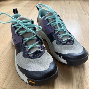 May include: A pair of athletic sneakers featuring a light gray mesh upper with dark gray overlays, purple accents, and bright turquoise laces. The shoes have a thick white midsole and a black rubber outsole with a yellow Vibram logo on the side.