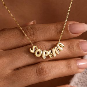 May include: A gold-toned necklace featuring a delicate chain and a central pendant composed of individual, rounded, three-dimensional letters that spell out the name Sophia in a polished, metallic finish that reflects light.