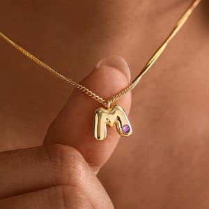 May include: A gold-toned chain necklace featuring a small, puffed letter M pendant. The pendant has a smooth, polished finish and is accented with a single, small, round purple gemstone embedded on the right side of the letter's curve.