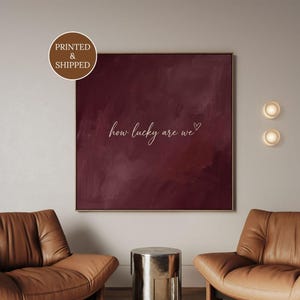 May include: A square wall art piece featuring a deep burgundy, brushstroke-textured background with the white cursive text "how lucky are we" followed by a small heart symbol. The artwork is framed in a thin, neutral-toned border.