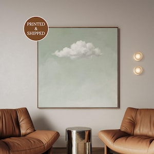 Puede incluir: Una pieza de arte mural cuadrada que presenta una sola nube blanca centrada sobre un fondo verde salvia apagado, enmarcada con un borde metálico fino. La obra incluye una insignia redonda marrón con el texto PRINTED & SHIPPED en blanco.