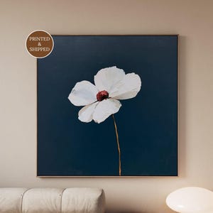 May include: A square wall art print featuring a single white flower with a dark red center against a solid navy blue background. The flower has thick, textured petals and a thin, light brown stem. A circular badge in the corner reads PRINTED & SHIPPED.